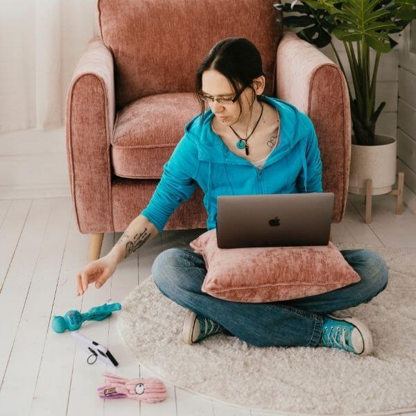 Jeni sitting on a rug with a laptop on a pillow, reaching for her phone, surrounded by dog toys in a cozy room.