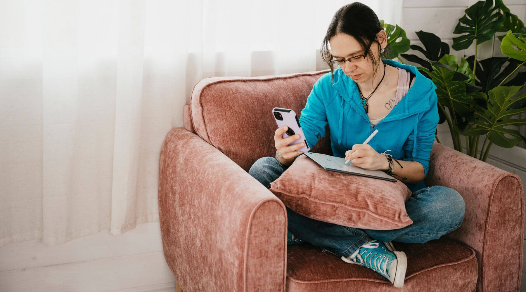 Jeni sitting cross legged in a blue hoodie, on a pink armchair using a phone and writing in a notebook.