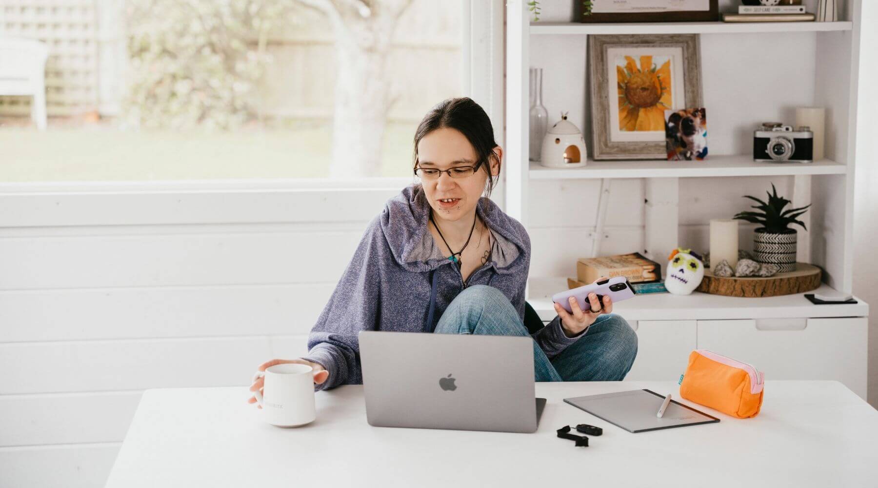 Jeni sitting at a desk with a laptop, phone, and coffee mug in a home office setting.